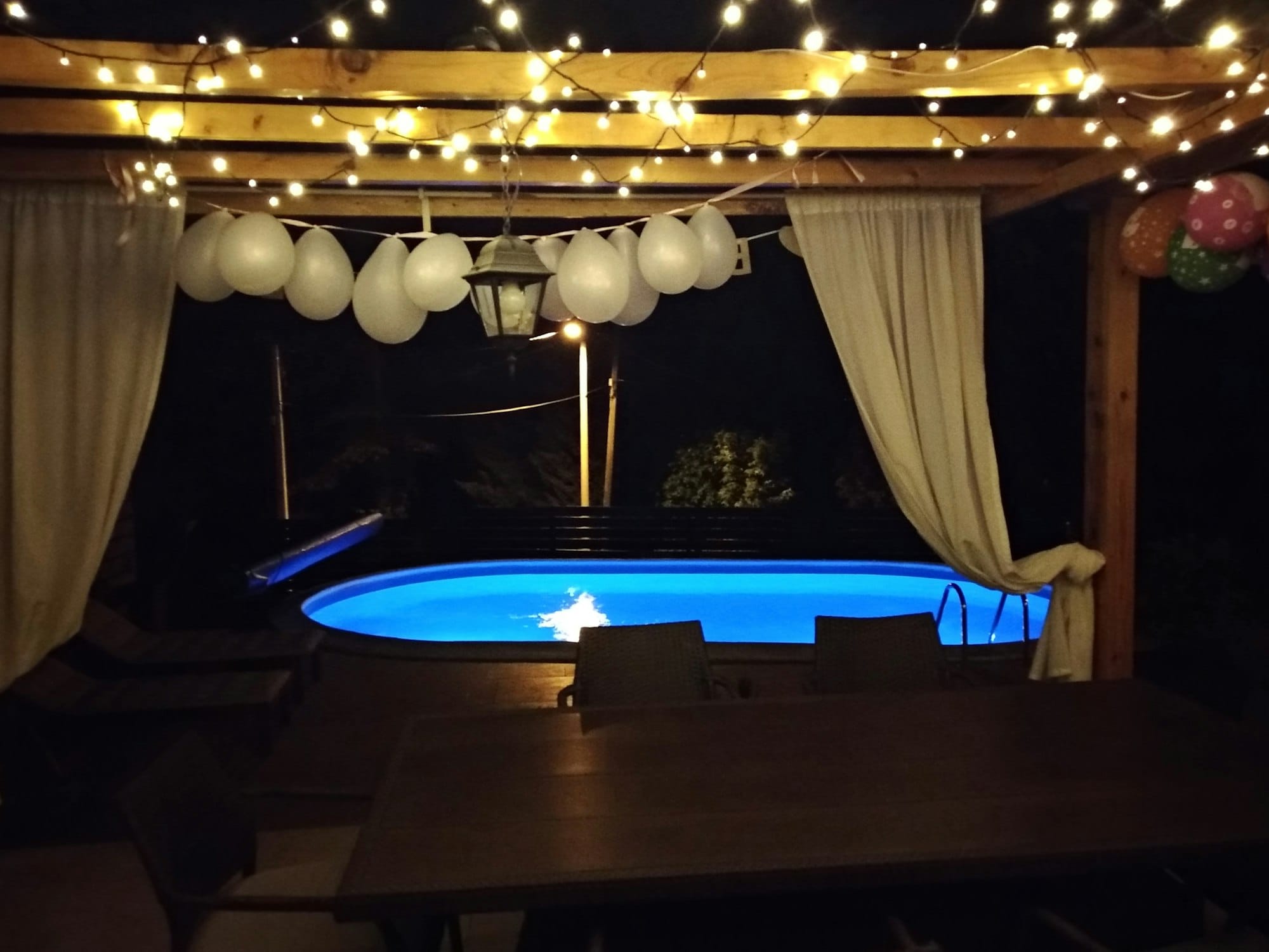 A blue swimming pool in front of a terrace with lovely evening lights and baloons.