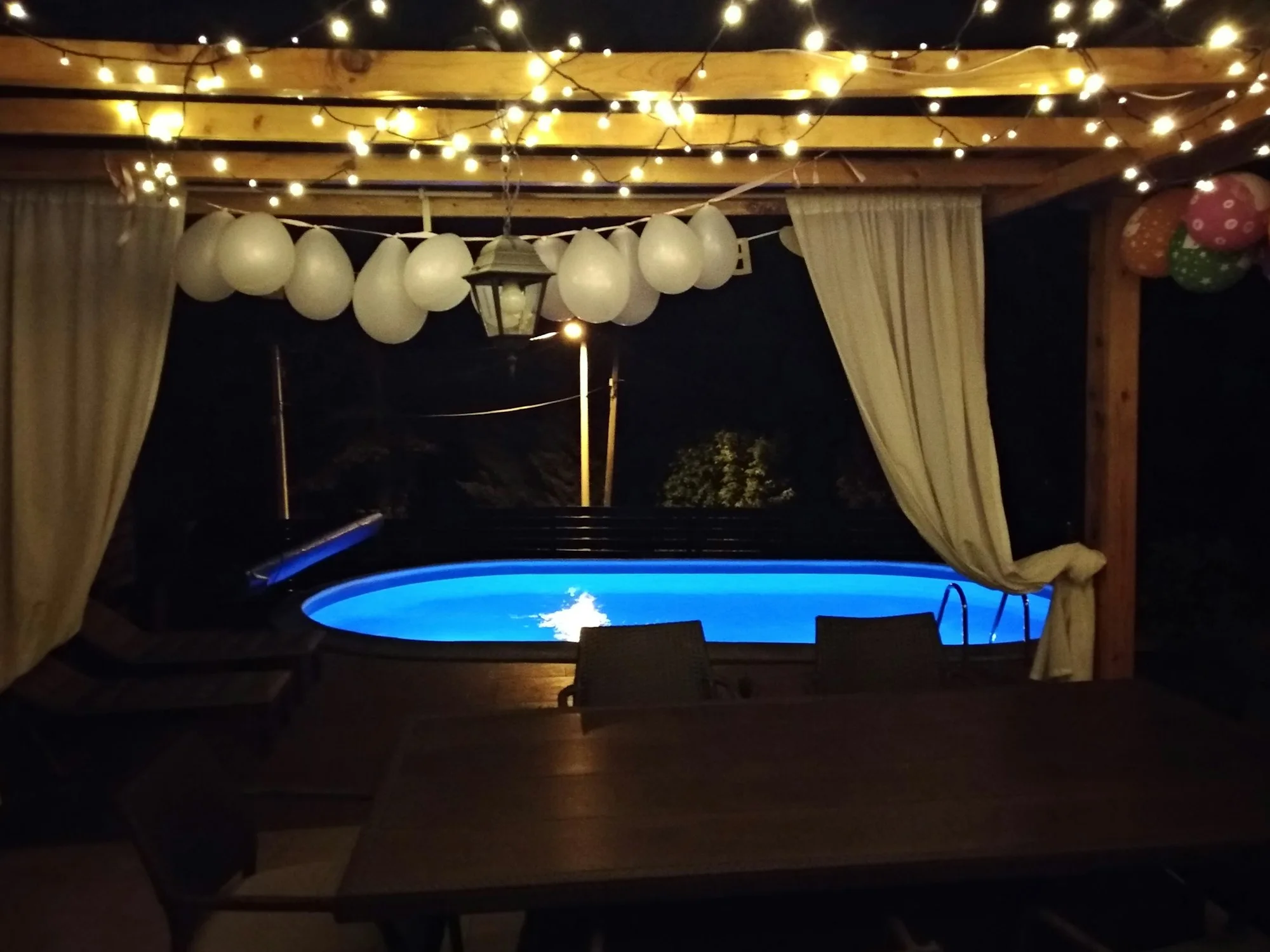 A blue swimming pool in front of a terrace with lovely evening lights and baloons.